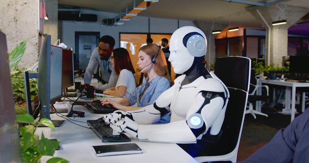 Office with long desk with computers. A robot is sat typing in the foreground, with three humans behind it.